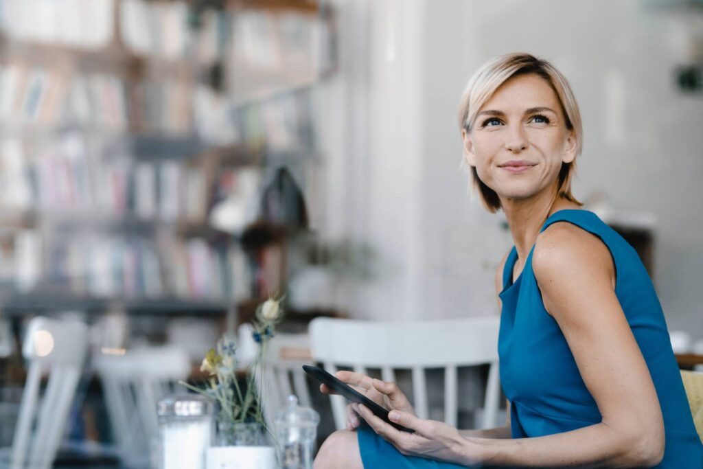 Selbstbewusste Frau in einem modernen Café oder Coworking-Space nutzt ein Tablet. Im Hintergrund sind Bücherregale und ein stilvolles Ambiente zu erkennen. Die Szene steht für digitale Selbstbestimmung, inklusive Arbeitskultur, digitale Teilhabe und moderne Arbeitsplatzgestaltung. Ideal zur Darstellung von Frauen in der digitalen Arbeitswelt und barrierefreiem Arbeiten unterwegs.