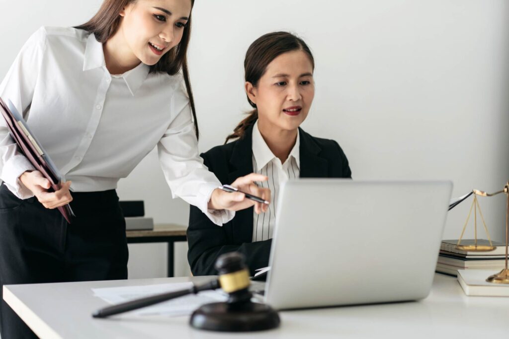 wei Juristinnen arbeiten konzentriert an einem Laptop in einem modernen Büro. Eine der beiden zeigt auf den Bildschirm, während ein Richterhammer auf dem Tisch die juristische Arbeitsumgebung unterstreicht. Die Szene steht exemplarisch für digitale Barrierefreiheit in der Rechtsberatung, barrierefreie Gesetzgebung und die Umsetzung von Accessibility-Standards im E-Government.
