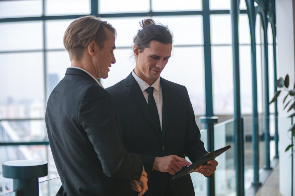 Zwei Geschäftsmänner in Anzügen stehen in einem modernen Bürogebäude mit Glasfront und blicken gemeinsam auf ein Tablet. Die Szene vermittelt professionelle Zusammenarbeit, strategische Planung und digitale Transformation im Business-Umfeld. Keywords wie digitale Barrierefreiheit, inklusive Unternehmensstrategie, digitale Teilhabe, barrierefreie Kommunikation und Leadership in der digitalen Arbeitswelt sind relevant.