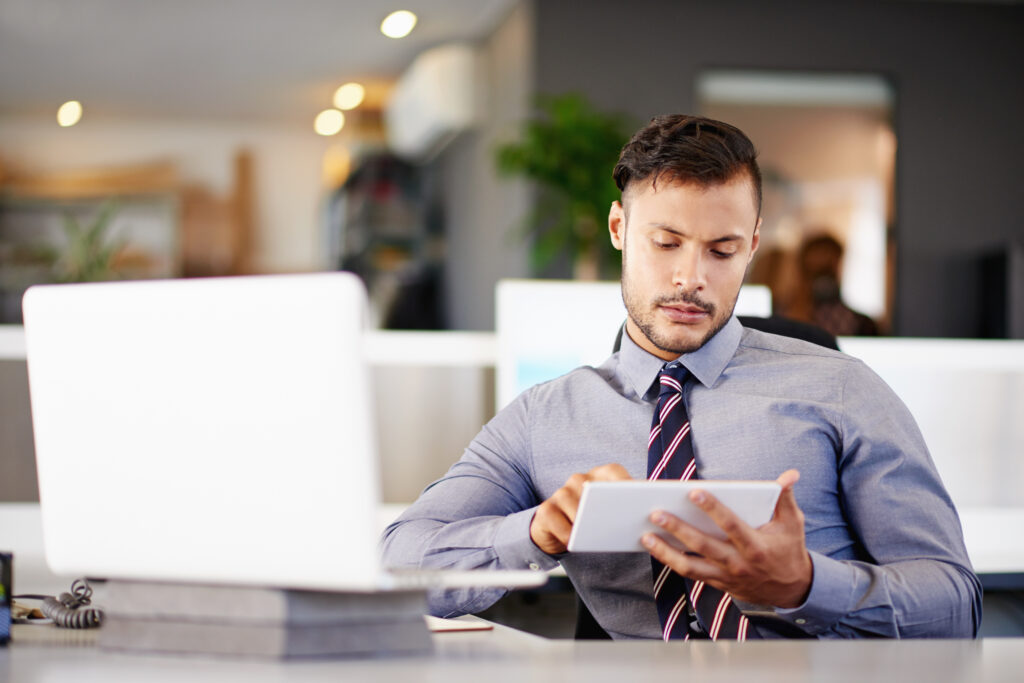 Ein junger Mann im Hemd und Krawatte sitzt in einem modernen Büro vor einem Laptop und schaut konzentriert auf ein Tablet. Die Szene steht sinnbildlich für digitale Verwaltungsprozesse und zeigt, wie barrierefreie E-Government-Dienste und digitale Arbeitsplätze zunehmend mobil genutzt werden können.