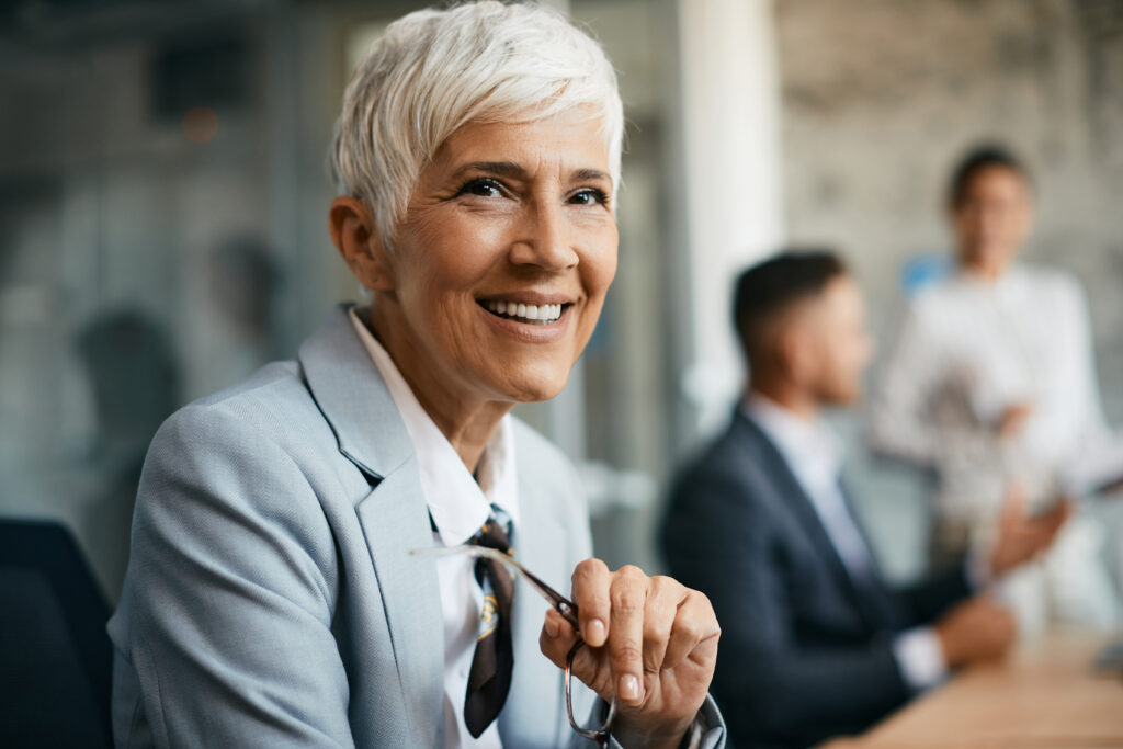 Eine ältere Geschäftsfrau mit kurzen weißen Haaren blickt selbstbewusst in die Kamera. Sie sitzt in einem modernen Büro, im Hintergrund sind Kolleg:innen in einem Meeting zu sehen. Die Szene steht für digitale Inklusion, barrierefreie Teilhabe im Berufsleben und Diversity am Arbeitsplatz – besonders in Führungspositionen. Ein Beispiel für Senior Leadership in einem inklusiven Arbeitsumfeld, das digitale Barrierefreiheit fördert.
